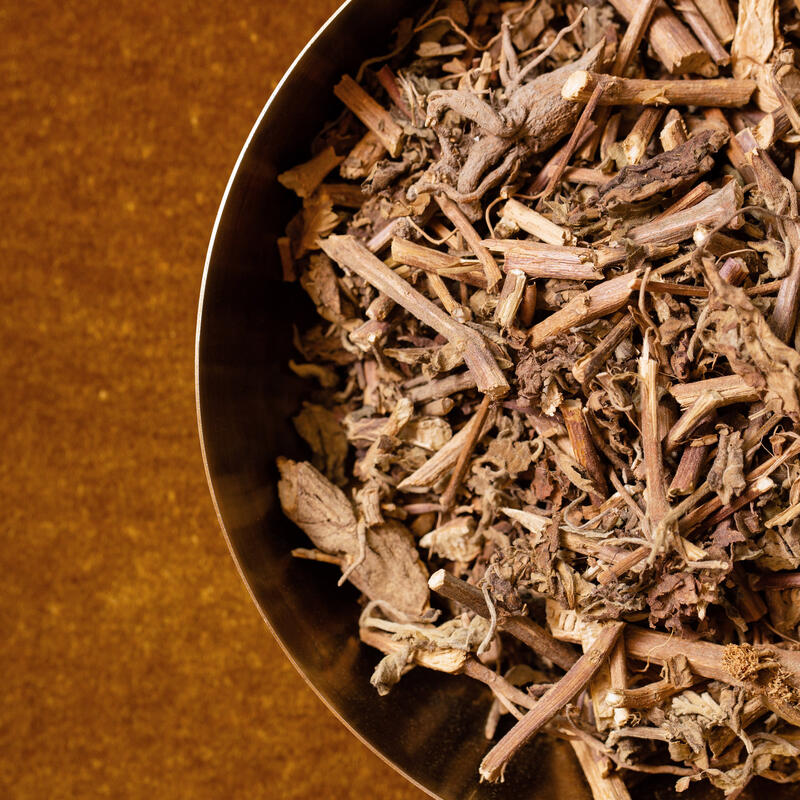 Dried tea leaves and twig fragments fill a round metal bowl, resting on a warm brown textured surface.