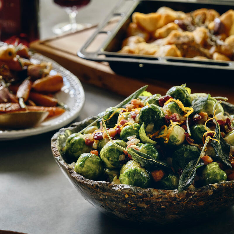 Bowl of roasted Brussels sprouts garnished with herbs and nuts, set on a dining table with other dishes including roasted carrots and a pastry dish, surrounded by wine glasses.