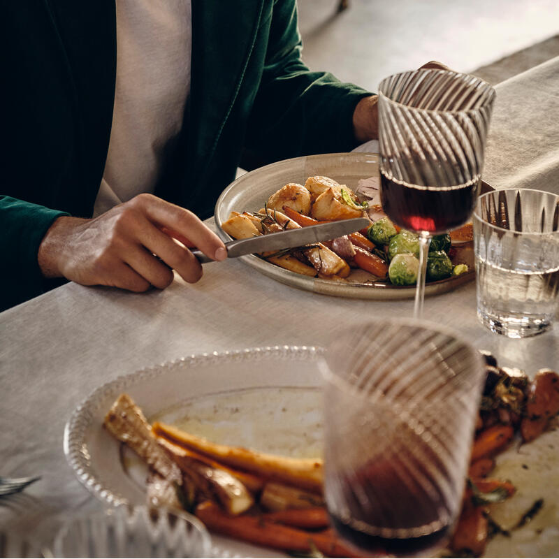 A person in a green jacket cuts roasted vegetables on a plate at a white table, surrounded by wine glasses and additional dishes in a warmly lit room.