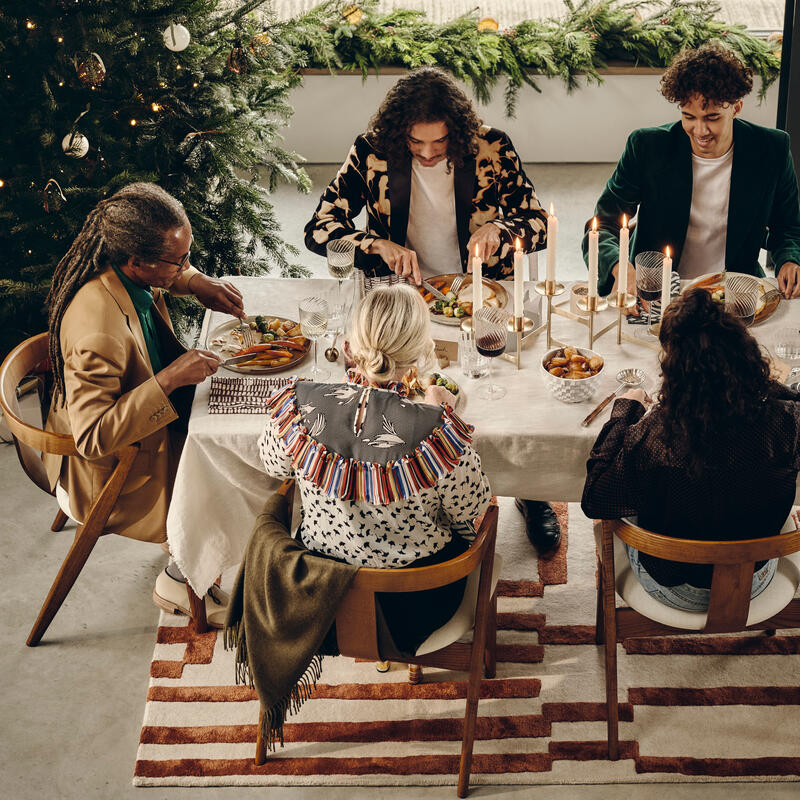 Five people sit around a festive dinner table with candles and wine glasses, sharing a meal. Behind them is a decorated Christmas tree and greenery on the window ledge.