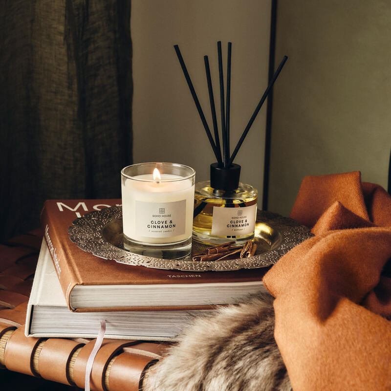 A lit "SOHO HOUSE CLOVE & CINNAMON" candle and diffusers rest on a tray atop two stacked books, surrounded by a brown scarf and fur throw on a woven leather seat.