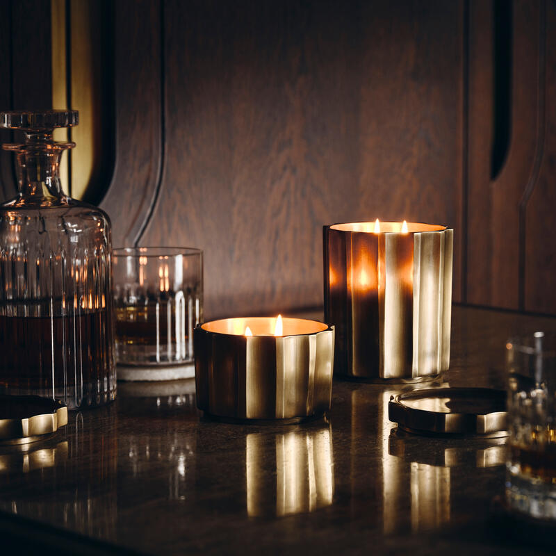 Two golden candles are burning on a reflective surface next to crystal glassware, including a decanter and a whiskey glass, in a dimly lit wooden-paneled room.