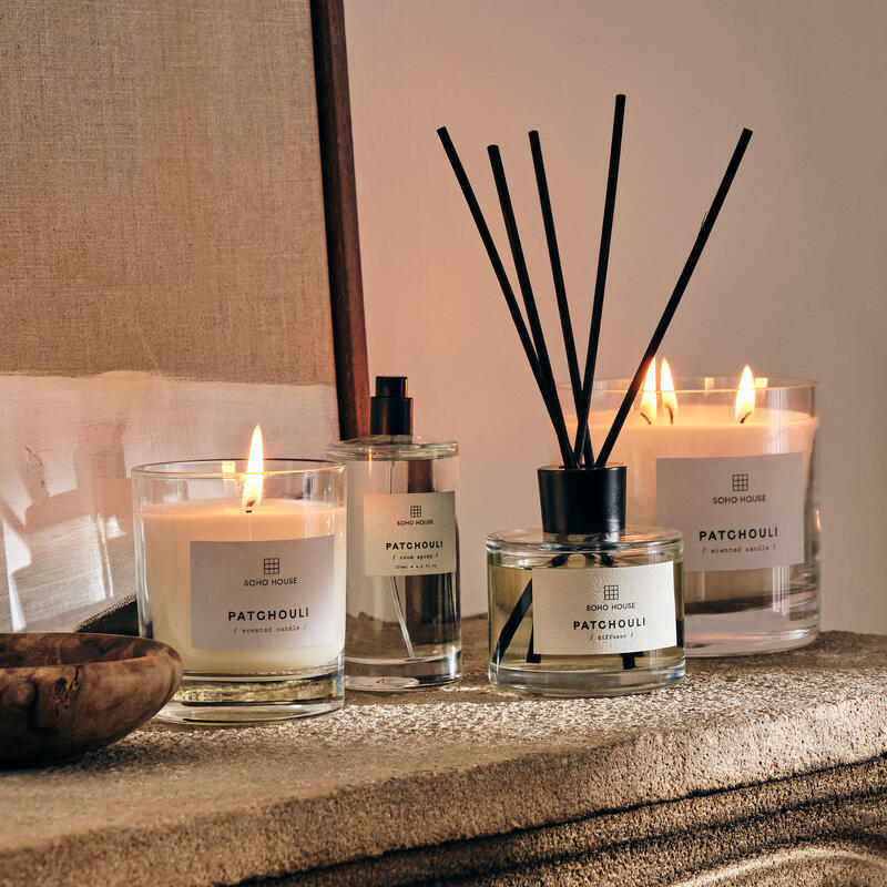 Candles and a diffuser labeled "SOHO HOUSE PATCHOULI" sit on a textured stone shelf beside wooden and linen decor, softly illuminated by candle flames casting warm light.