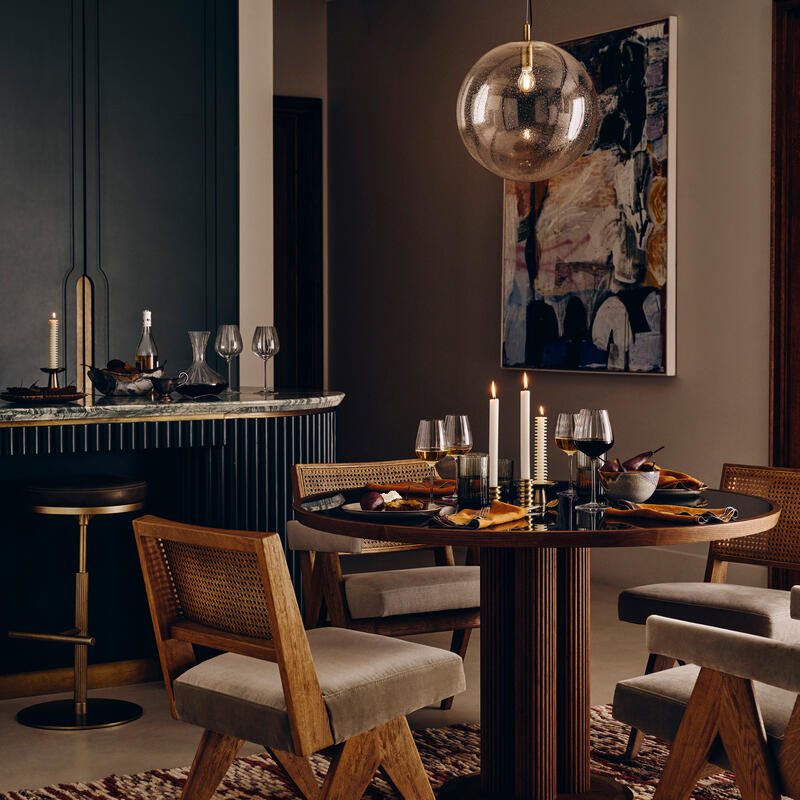 A round dining table set with candles and glassware is surrounded by four cushioned wooden chairs on a patterned rug, under a spherical pendant light in a stylish, modern dining area.