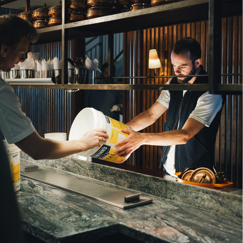 Two men in a kitchen transfer contents of a large Hellmann’s Real Mayonnaise container, surrounded by stacked pots and tableware on marbled countertops, under warm indoor lighting.