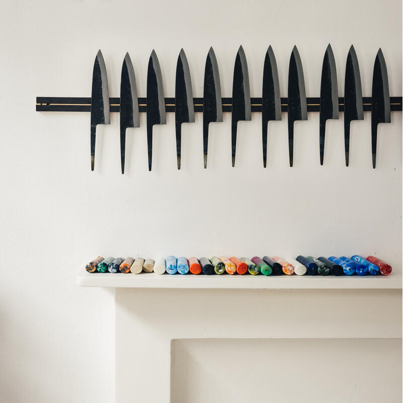 Knives are neatly aligned in rows on wall-mounted magnetic strips, while colorful crayons are arranged on a white shelf below, against a plain white wall.