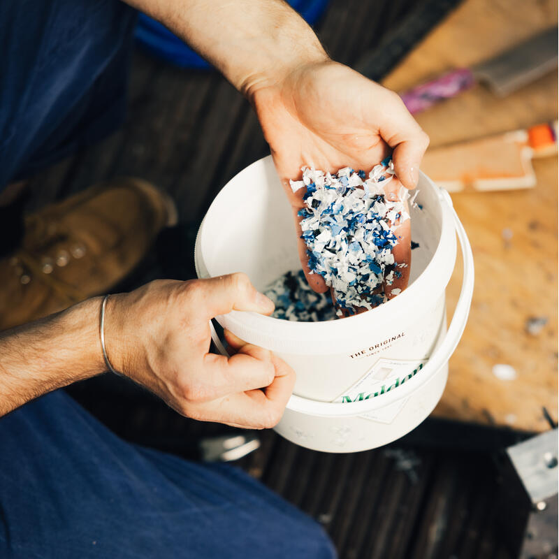 Hands hold a plastic bucket filled with blue and white shredded material, positioned against a wooden surface with tools and equipment around. Text on the bucket reads, "THE ORIGINAL."