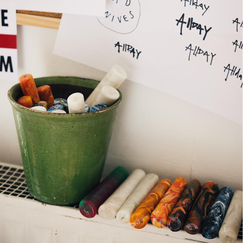 Colored wax sticks are placed in a green pot and lined up on a white surface. In the background, sheets with various "ALL DAY" handwriting styles are pinned to a corkboard.