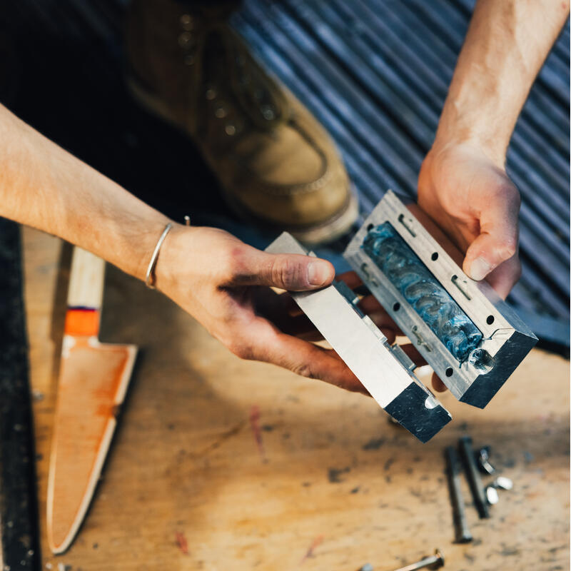 Hands holding a metal mold with blue residue, with a wooden-handled putty knife, screws, and metal shavings scattered on a wooden surface; a booted foot is partially visible in the background.