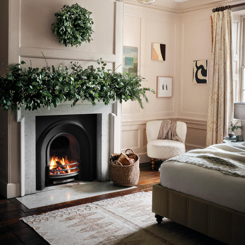 A marble fireplace with a roaring fire is adorned with greenery, situated in an elegantly decorated bedroom with framed art, a wicker basket, and a modern chandelier overhead.