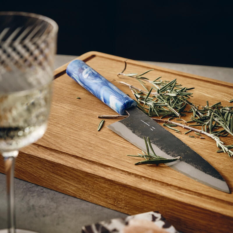 A knife with a blue handle lays on a wooden cutting board among scattered rosemary, set against a dark background, with a glass of white wine and a rock salt block nearby.