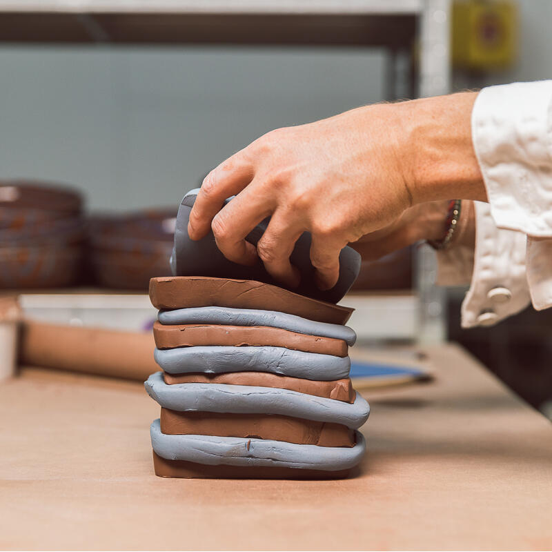 A hand arranges a stack of alternating brown and gray clay slabs on a worktable, with shelves holding finished pottery in the background.