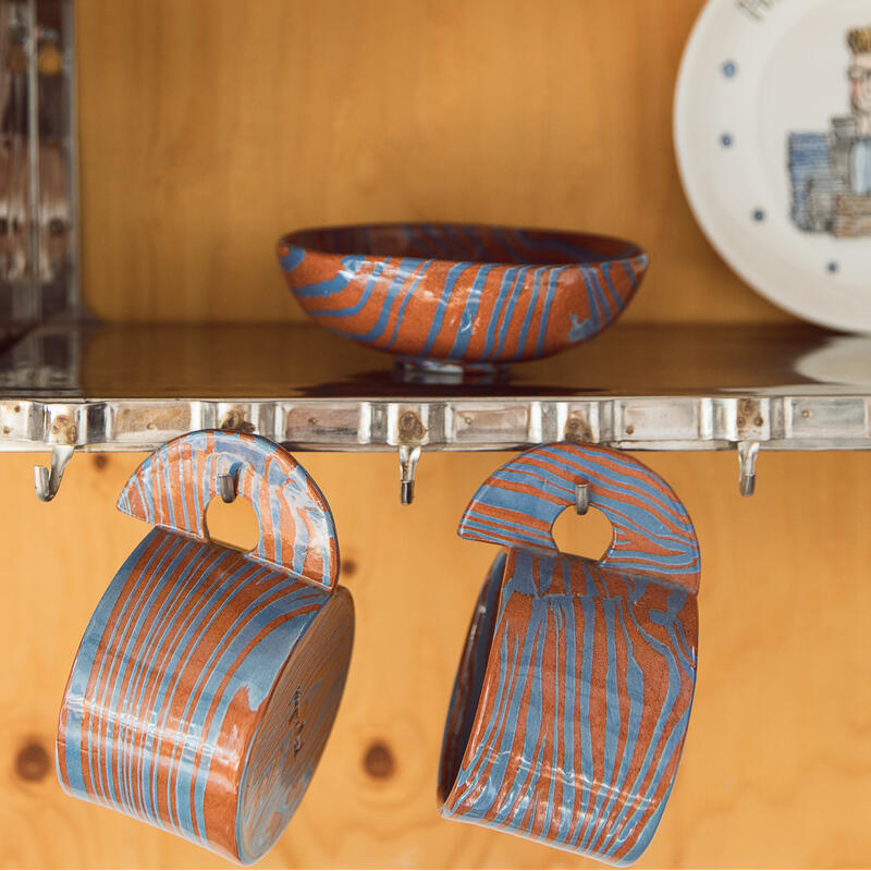 Colorful ceramic plates, bowls, and cups are drying on a stainless steel rack mounted on a wooden wall. Plates are aligned vertically, a bowl lies flat, and cups hang below.