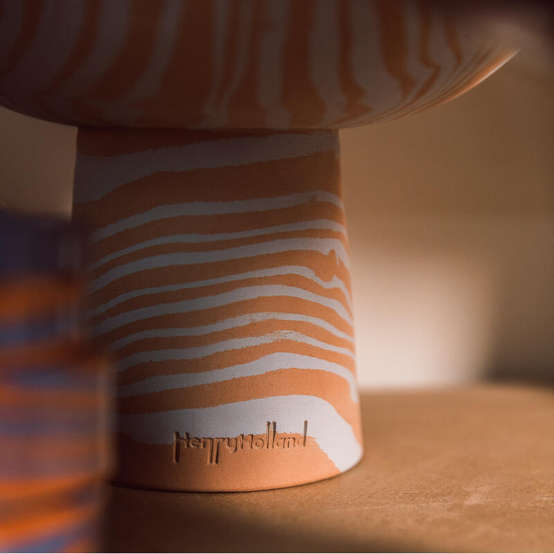 Ceramic vase with white and orange wavy stripes sits on a wooden surface in soft light. “HenryHolland” is etched at the base. Another blurred striped object is in the foreground.