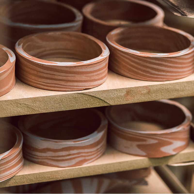 Several circular clay pots are drying on wooden shelves within a pottery workshop, showcasing their natural, unfinished state. The pots display marbled patterns, highlighting the artisan's craftsmanship.