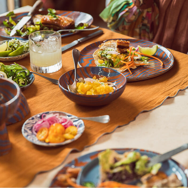 Plates with colorful salads, a glass of a white beverage with ice, and various garnishes on a table set with an orange cloth, located in a cozy dining setting.