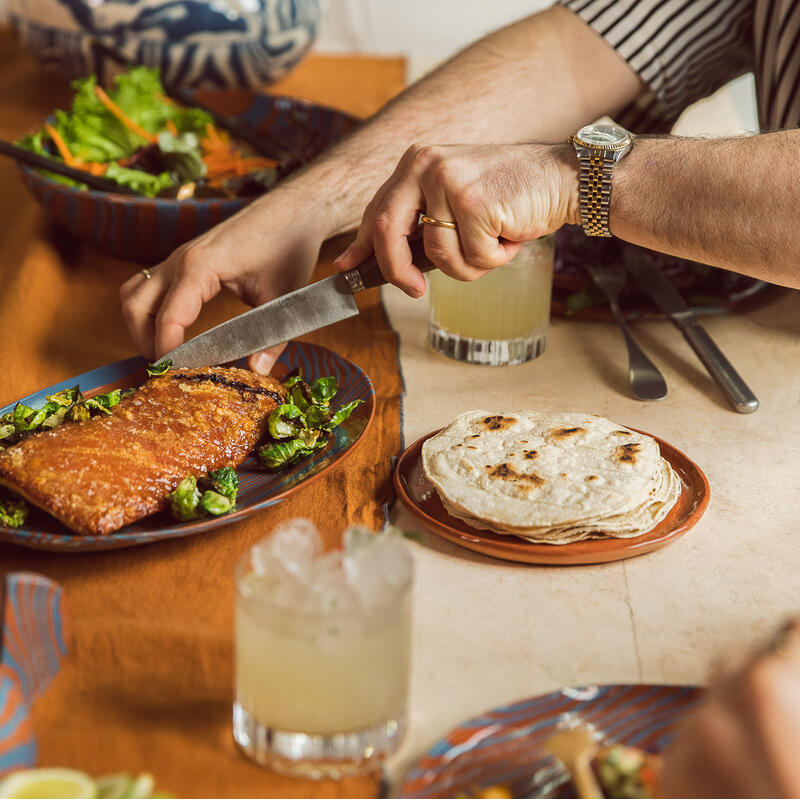 Man cutting crispy glazed pork belly on plate, surrounded by other dishes including tortillas, salad, lime wedges, and drinks. The setting features a decorative blue vase with plants on a tan table.