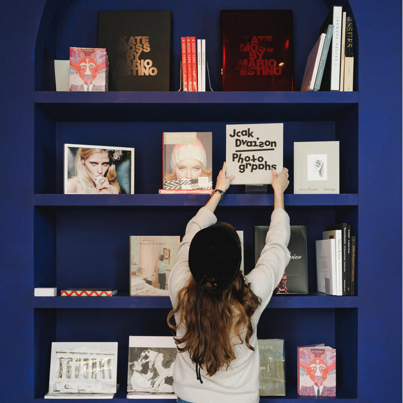 A person reaches up to arrange a book titled "Jack Davison Photographs" on a blue bookshelf filled with various books and photo albums within a modern interior space.