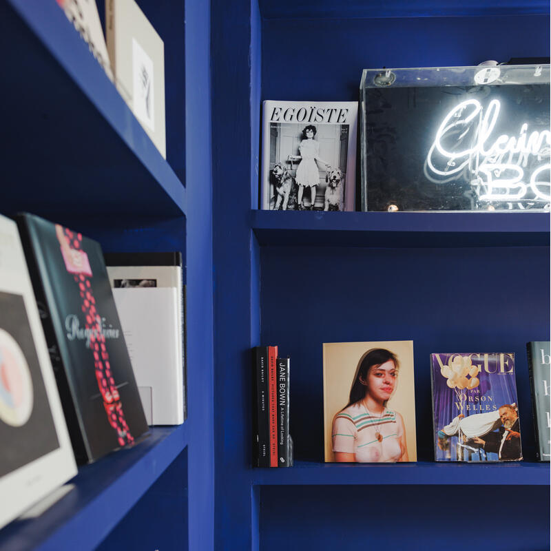 Books are arranged on blue shelves; one shelf displays a magazine titled "EGOISTE". Below are more books and a photo. Yellow cushion and patterned pillow lie nearby.