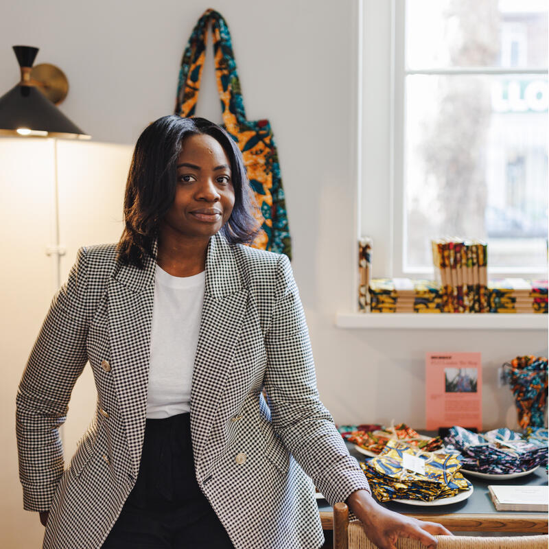 A woman stands confidently in a checkered blazer and white shirt, leaning on a table with vibrant, patterned fabrics in a well-lit room.