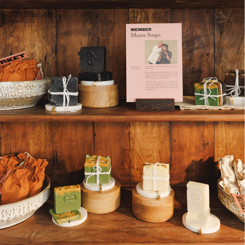 Handmade soaps are arranged on wooden shelves, surrounded by vases and bowls of various textures. A pink display card reads: "MEMBER Mums Soaps. Mums Soaps is a... the way it should be lived."