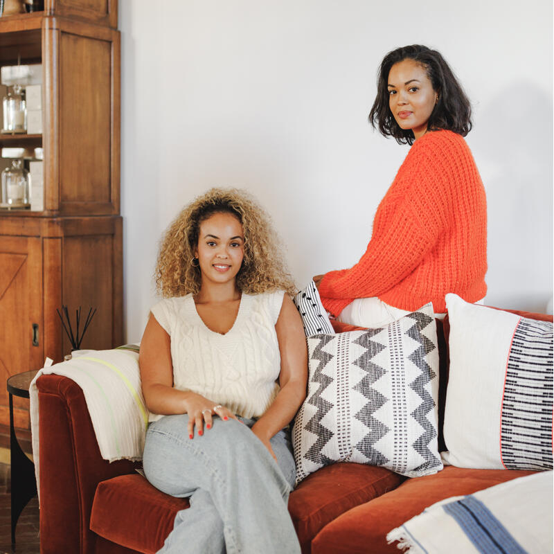 Two women sit on a red sofa adorned with various patterned pillows; one leans back, the other sits upright in a cozy living room with wooden shelves displaying décor items.