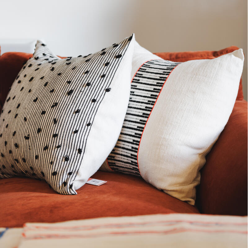 Two decorative pillows, one with black dots on stripes and the other with a striped band, rest on an orange couch in a minimalistic, well-lit room.