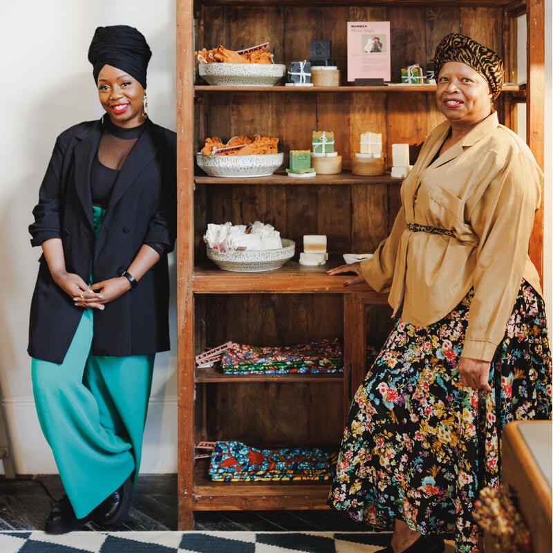 Two women stand beside a wooden shelf holding various items including baskets, jars, soaps, and fabrics, in a brightly lit room with a patterned floor.