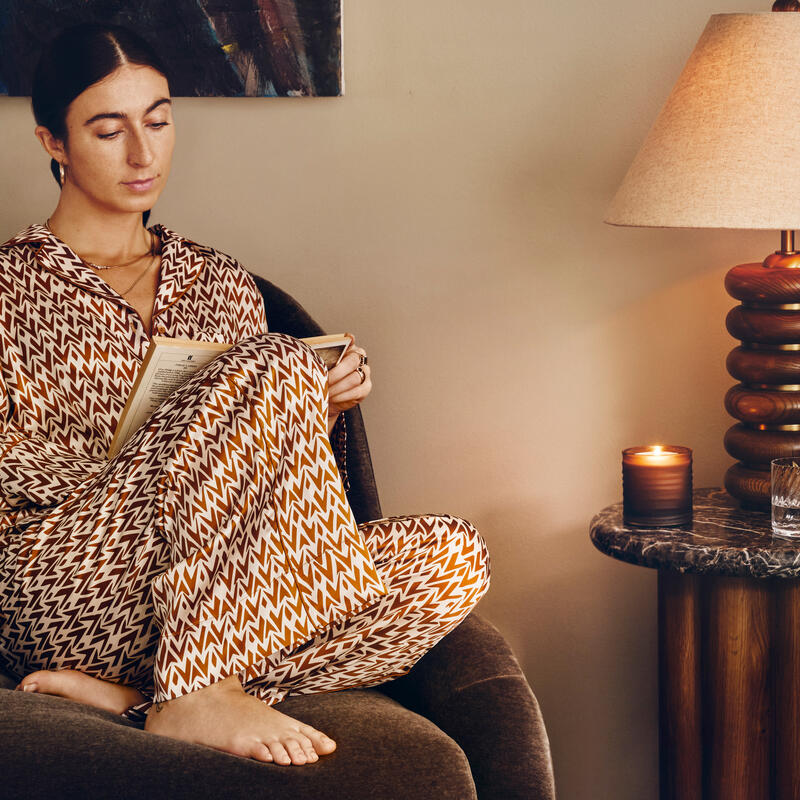 A person sits in a chair, reading a book while wearing a geometric pattern outfit. A lit candle and lamp sit on a small side table. A colorful painting hangs behind.