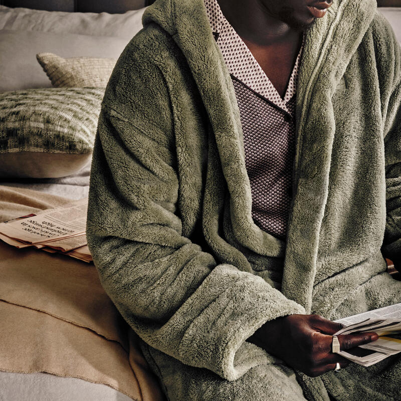 Person wearing a textured, light green robe and patterned pajama set, sits on a plush, layered bed, holding open papers in both hands, with a scattered newspaper in the background.