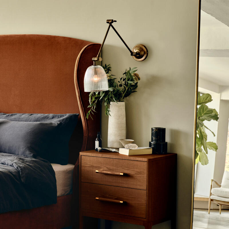 A wooden nightstand with a book, vase of greenery, and a lamp; positioned next to a brown upholstered bed and a large mirror, adjacent to a cozy living area with plants.