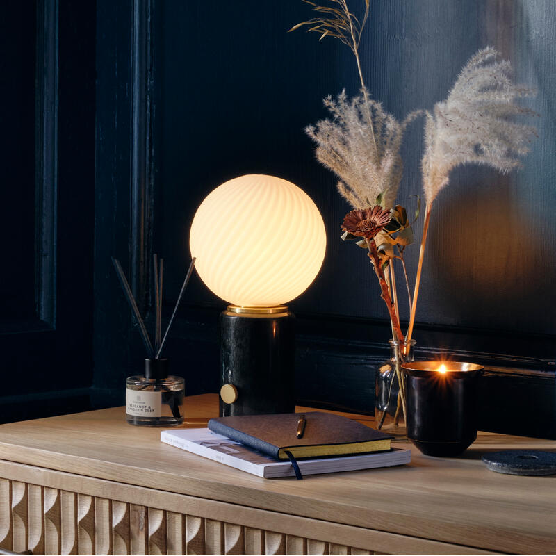 A glowing spherical lamp stands on a wooden dresser with textured drawers, surrounded by books, a candle, a vase of dried flowers, and incense in a dark-walled room.