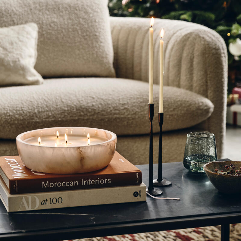 A bowl candle with multiple lit wicks and two tall candles sit on a coffee table beside books titled "Moroccan Interiors" and "AD at 100" in a cozy living room with a decorated Christmas tree.