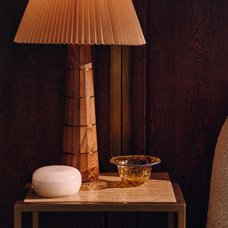 A wooden table lamp with a pleated shade sits on a small side table beside a white decorative item and a speckled glass bowl, with a textured armchair and dark wooden paneling in the background.