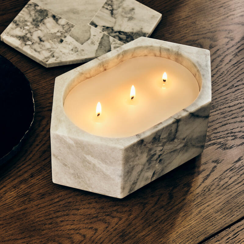 A rectangular marble container holding a lit three-wick candle sits on a wooden table, next to hexagonal marble coasters.