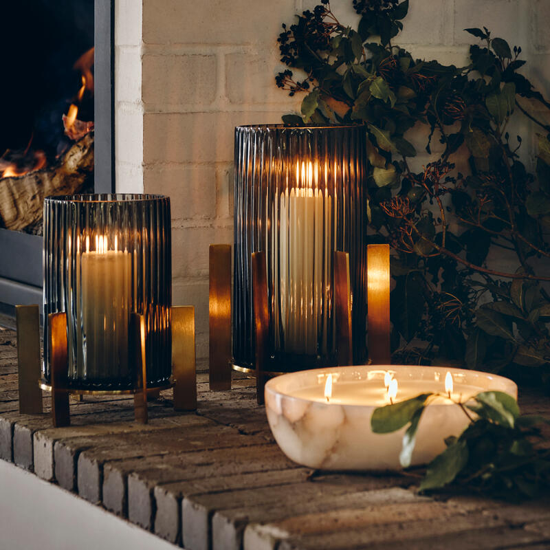 Candles burning in glass holders and a marble bowl on a brick mantel, with a fireplace on the side and leaves as decoration.