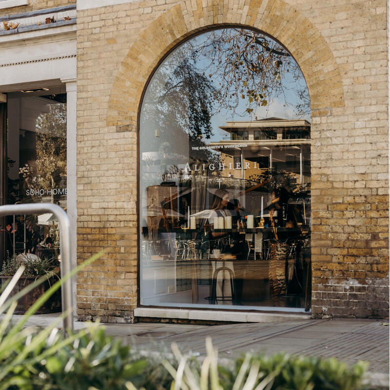 Arched window reflecting trees and buildings; inside, a store with “THE GOLDSMITH'S WORKBENCH” and “ALIGHIERI” signage. Brick exterior, neighboring “SOHO HOME” shop, surrounded by urban greenery.