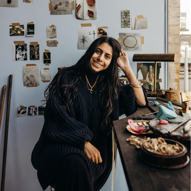 A woman sits smiling at a cluttered desk surrounded by art supplies and pinned sketches, against a softly lit wall with taped photographs and drawings, beside a window.