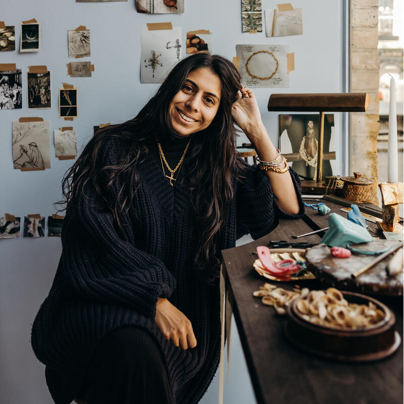 A woman, smiling and seated in a cozy workspace, touches her hair. Artifacts and photos are taped on the wall behind her, while creative materials cover the adjacent wooden desk.