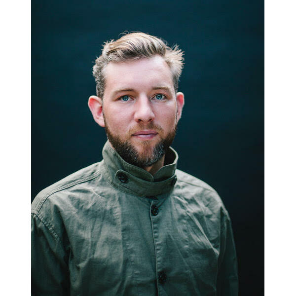 A person with short hair and a beard wears a high-collared, buttoned-up green jacket, standing against a dark, plain background.