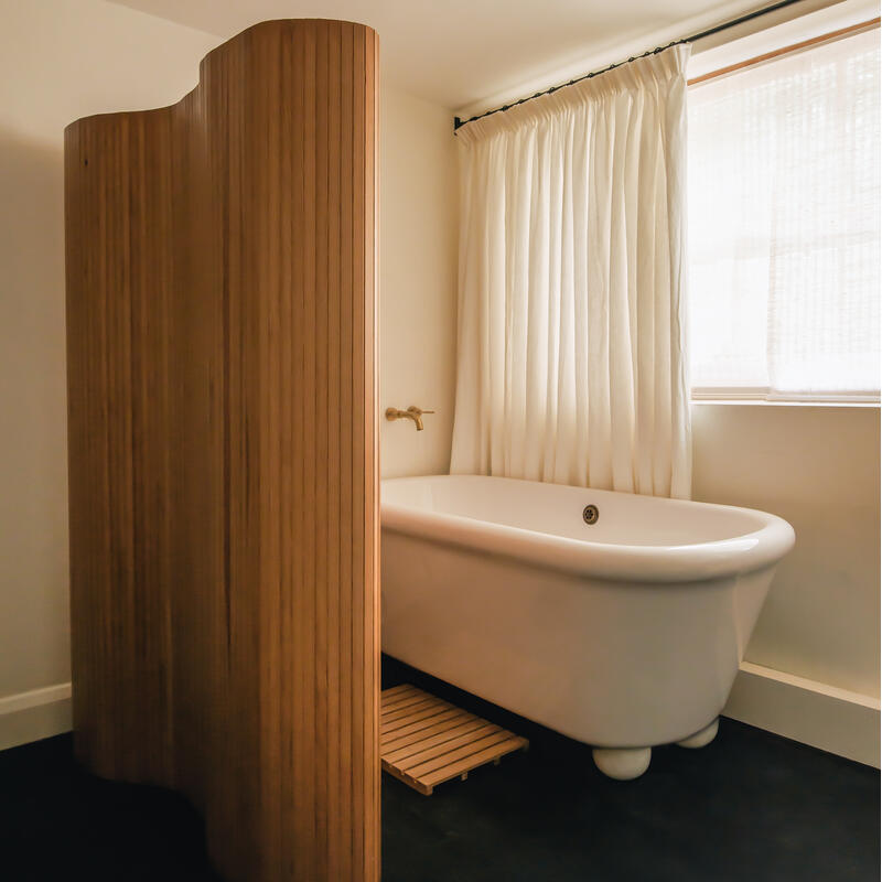 Freestanding bathtub with a white finish beside a wooden privacy screen in a minimalistic bathroom featuring beige walls, a dark floor, and a large window with white, translucent curtains.