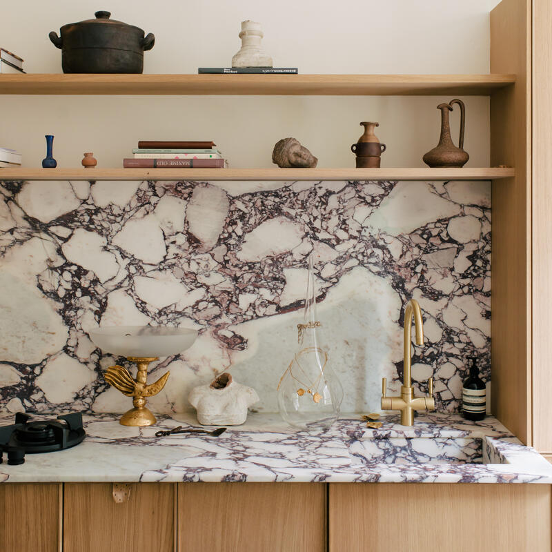 Marble countertop and backsplash with intricate patterns, featuring brass tap and sink. Wooden shelves above display pottery, books, and small sculptures in a compact, well-organized kitchen space.