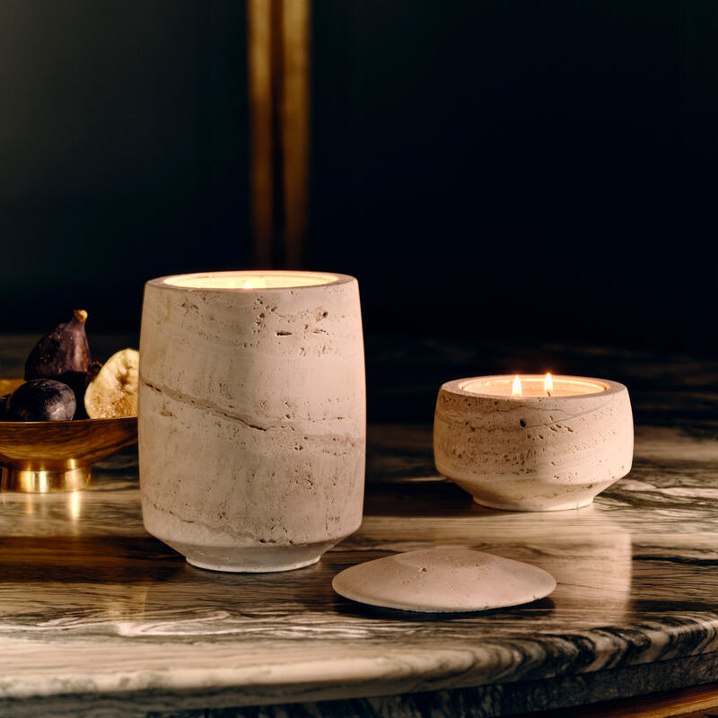 Ceramic candles glow softly on a polished marble surface; one smaller, one larger, next to a detached lid. A golden bowl with figs and slices of dried fruit completes the setting.