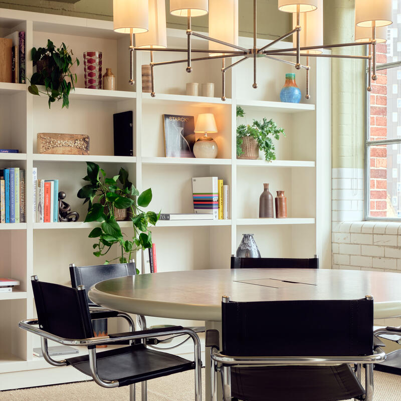 A round wooden table with black chairs is set in a room with white shelves filled with books, plants, and decorative items. Hanging above is a large metal chandelier with cylindrical lamps.