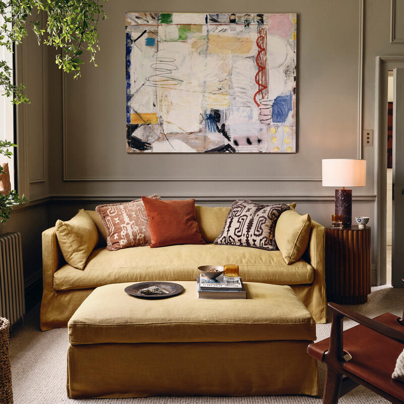 A mustard-yellow couch with patterned cushions sits in a cozy living room; a large abstract painting hangs above, surrounded by earthy-toned decor and a leafy green plant near a window.