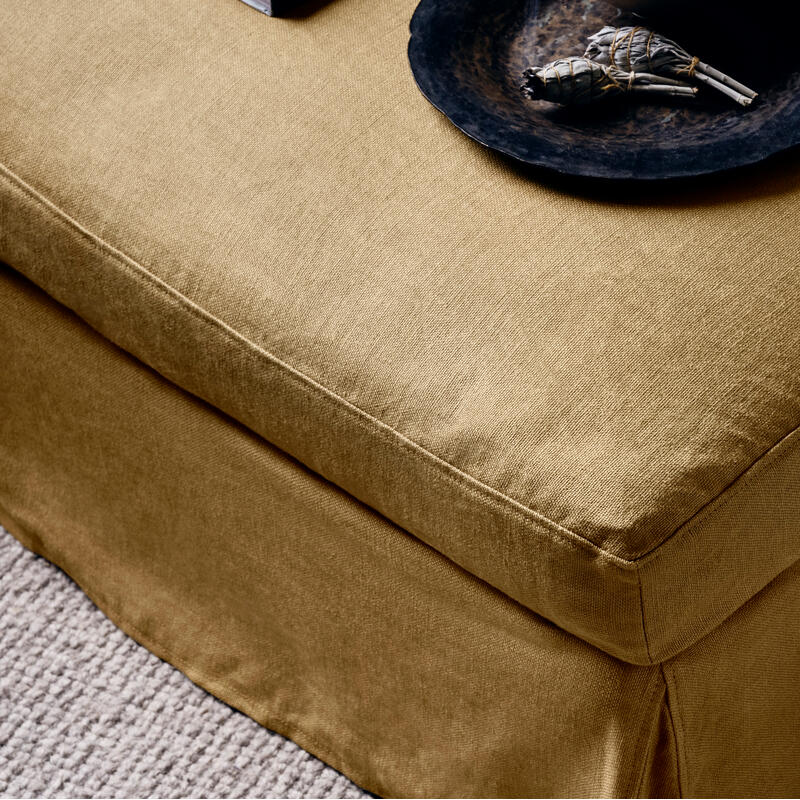 A beige upholstered ottoman holds a black bowl with a white sage bundle and potpourri, resting on a textured gray carpet. A closed book is partway visible.