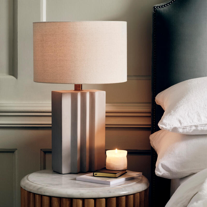 A light gray table lamp and lit candle rest on a round, marble-top nightstand with wooden panels, beside a bed with white linens and a headboard with nailhead trim in a softly-lit bedroom.