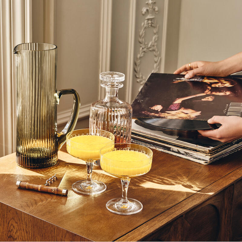 Two orange cocktails in elegant glasses sit on a wooden cabinet near a crystal decanter and a large pitcher. A hand flips through vinyl records nearby in a well-lit, ornate room.