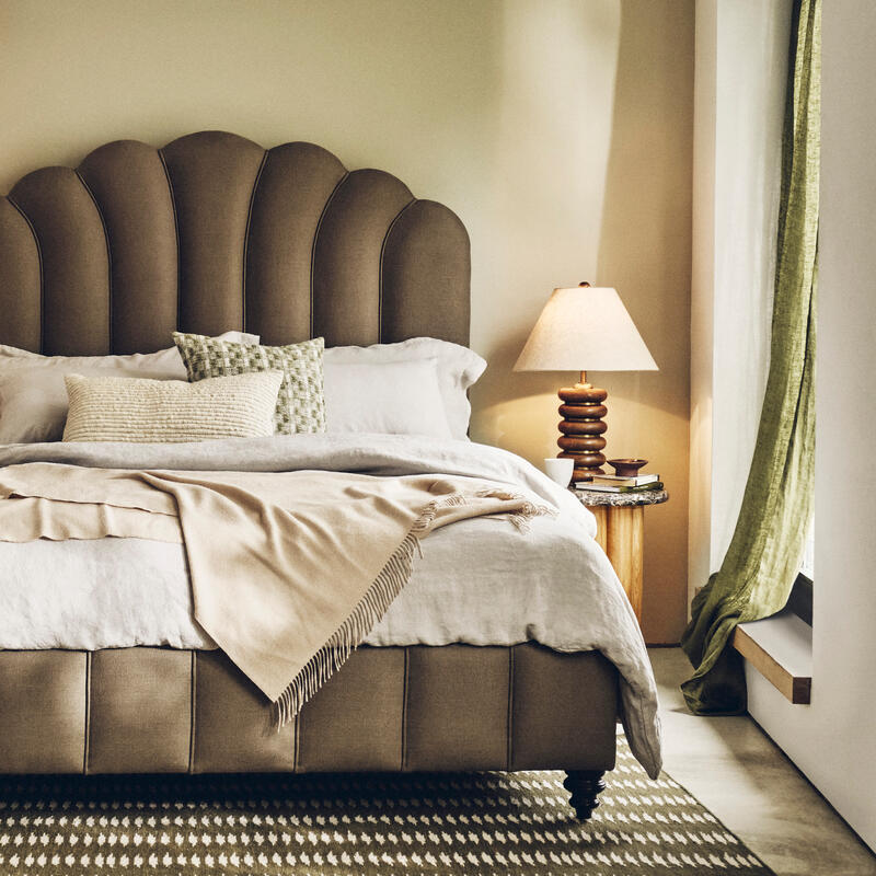 A neatly made bed with a scalloped headboard sits between two wooden nightstands holding lamps in a cozy, warmly-lit bedroom with beige walls, an armchair, patterned rug, and abstract wall art.