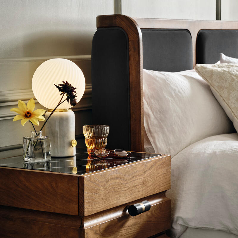 Wooden nightstand with a glowing lamp, a glass vase with flowers, glasses, and a glass, next to a bed with white bedding in a softly lit bedroom.